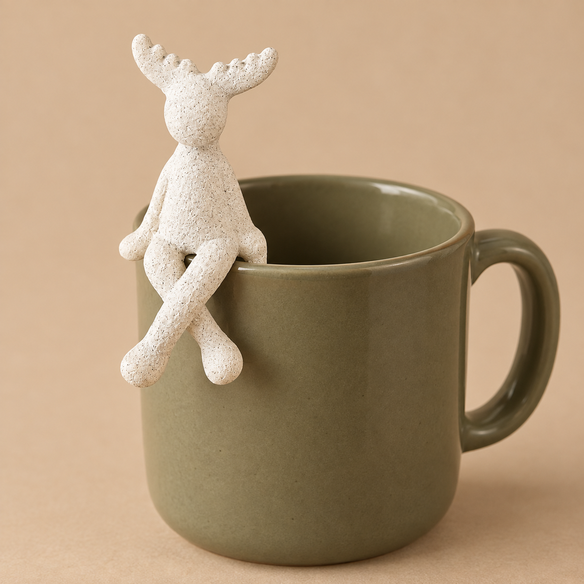 Kleiner weißer Elch Sören auf dem Rand einer olivfarbenen Tasse, mit außen herabhängenden Beinen