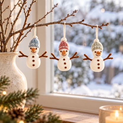 Drei Filz-Schneemänner Schneefreunde mit blauen, rosa und grünen Mützen an einem Zweig vor einem winterlichen Fenster.