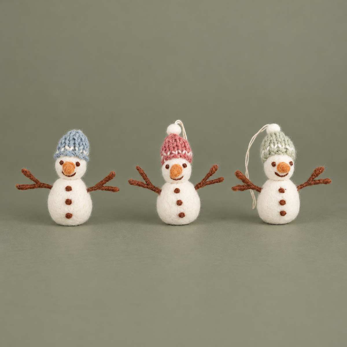 Drei Filz-Schneemänner Schneefreunde mit blauen, rosa und grünen Mützen vor gedecktem olivgrauem Hintergrund.